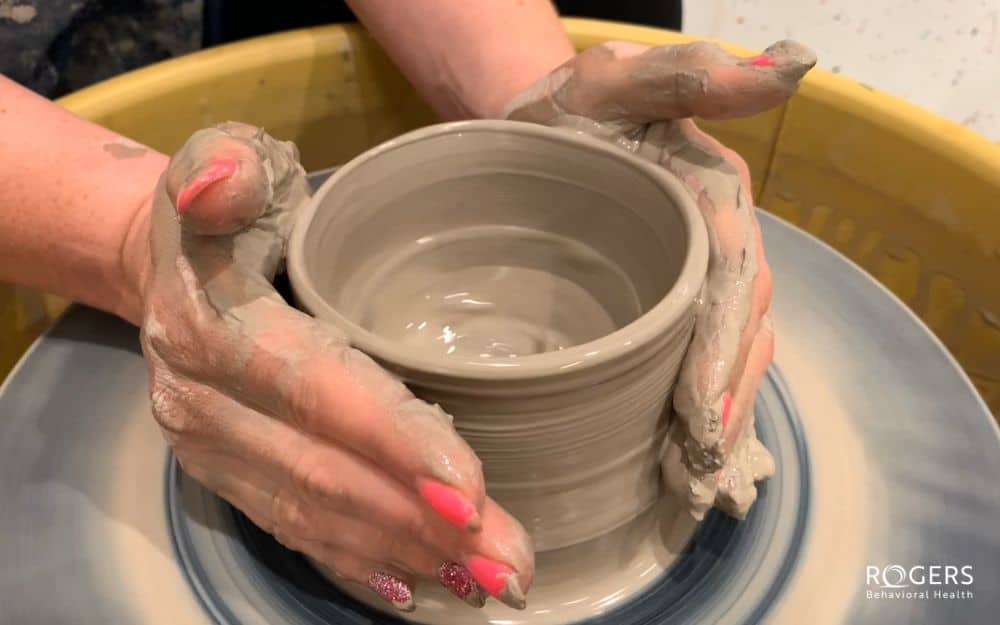 The image shows a close-up of two hands shaping a gray clay pot on a pottery wheel. The hands are coated in clay, and they have bright pink painted fingernails. The pottery wheel appears to be spinning, giving a sense of motion. The clay pot being formed is cylindrical, with concentric rings visible on its surface, indicating the rotation and shaping process. The background includes a blurred view of the edge of the pottery wheel, painted in a mix of blue and beige tones.