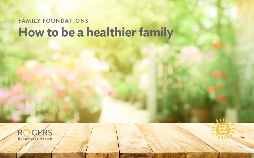 The image features a blurred background of a garden with lush greenery and colorful flowers in soft focus, creating a serene and vibrant atmosphere. In the foreground, there is a wooden table surface with visible grain and texture, adding a natural element to the image. The table is light brown with distinct planks. The text "FAMILY FOUNDATIONS How to be a healthier family" is displayed prominently at the top.