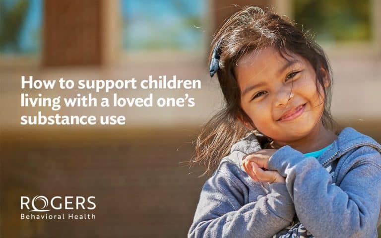 The image features a young girl outdoors, looking towards the camera with a gentle smile. She is wearing a blue-gray hoodie and a turquoise shirt underneath. Her dark hair is partially pulled back with a bow, and a few strands are blowing across her face. The background is softly blurred. On the left side of the image, there is white text : How to support children living with a loved one’s substance use