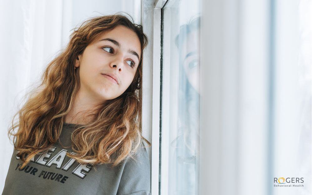 The image depicts a young person with long, wavy brown hair, wearing a gray sweatshirt with text. They are standing indoors, close to a window, and looking out. The image is bright, with the natural light coming through the window creating a soft illumination. The wall and window frame are painted white. The overall mood appears contemplative.