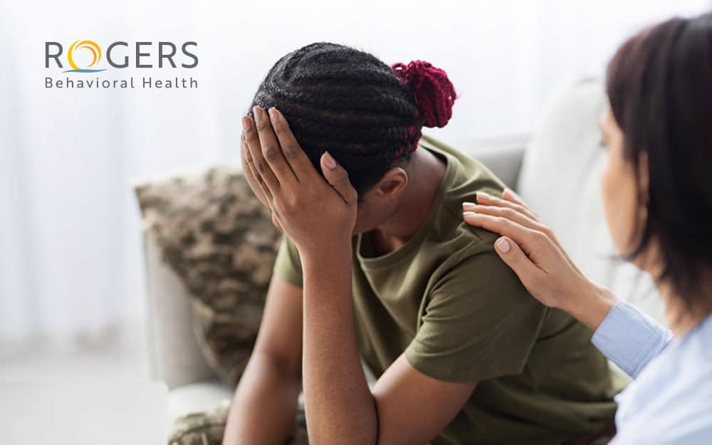 The image shows two individuals seated indoors, focusing on emotional support. The person on the left, with braided hair tied into a bun and wearing a green shirt, is leaning forward with a distressed expression, covering their face with one hand. The person on the right, wearing a light blue shirt, gently rests a hand on the other's shoulder in a comforting gesture. The background is softly blurred, featuring light-colored furniture and curtains.