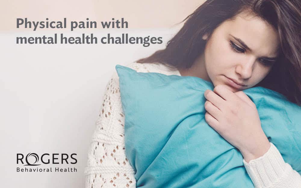 The image shows a young person with long dark hair, dressed in a white textured sweater, sitting and hugging a blue pillow. Their facial expression appears contemplative and somber. The background is a neutral light color, adding to the subdued mood of the image. In the upper left corner, there is text that reads, "Physical pain with mental health challenges".