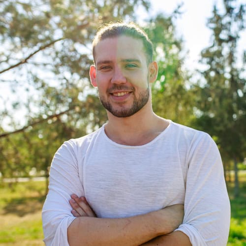A young adult male with tied-back brown hair and a beard smiles, standing in a sunlit park with trees in the background.
