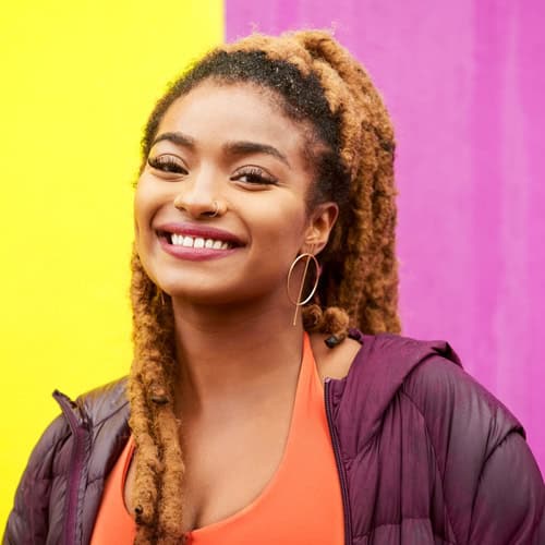 A person smiling in front of a yellow and pink background, wearing a purple jacket and orange top.