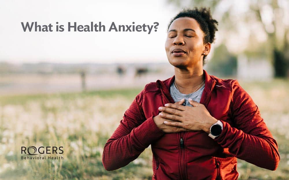 The image shows a person outdoors in a seated position, wearing a red jacket. They have their eyes closed and hands placed over their chest, appearing peaceful and meditative. The background is softly blurred with hints of greenery and distant structures, suggesting a natural setting. The sunlight casts a warm glow on the scene. In the upper left corner, text reads, "What is Health Anxiety?"
