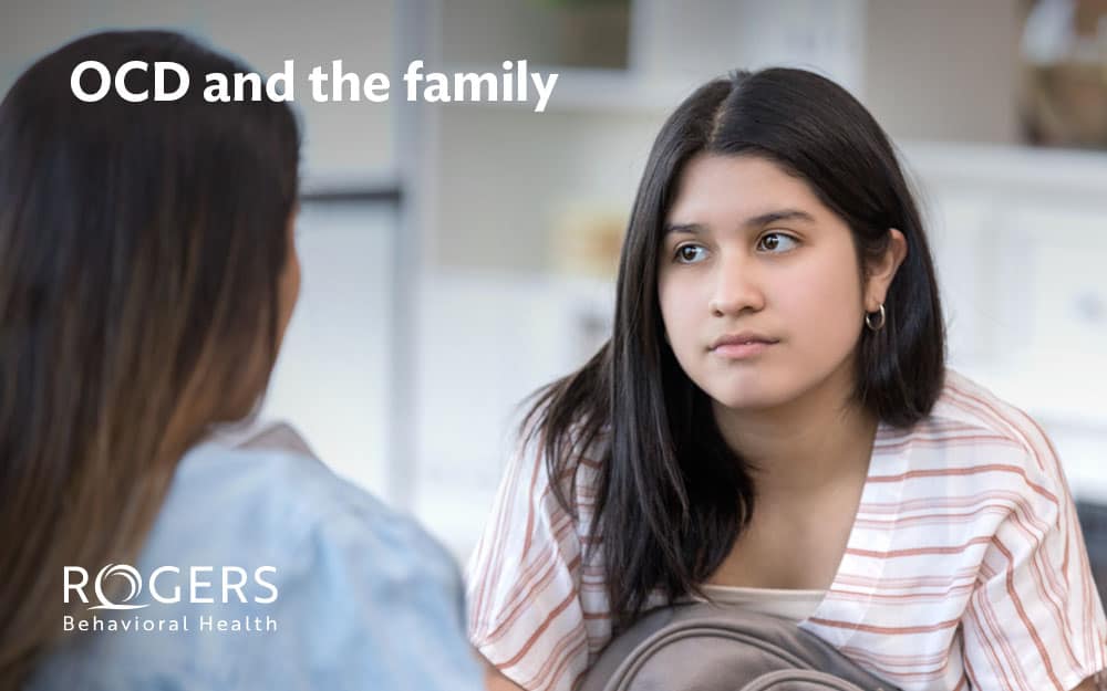The image portrays two individuals sitting indoors, engaged in what appears to be a serious conversation. The focus is on a young girl with long, dark hair, wearing a white and beige striped shirt, facing towards another person whose back is partially visible. This other person has long brown hair. The background is softly blurred, giving a sense of a cozy, home-like environment with white bookshelves and a soft color palette. Text overlayed at the top left corner reads "OCD and the family"