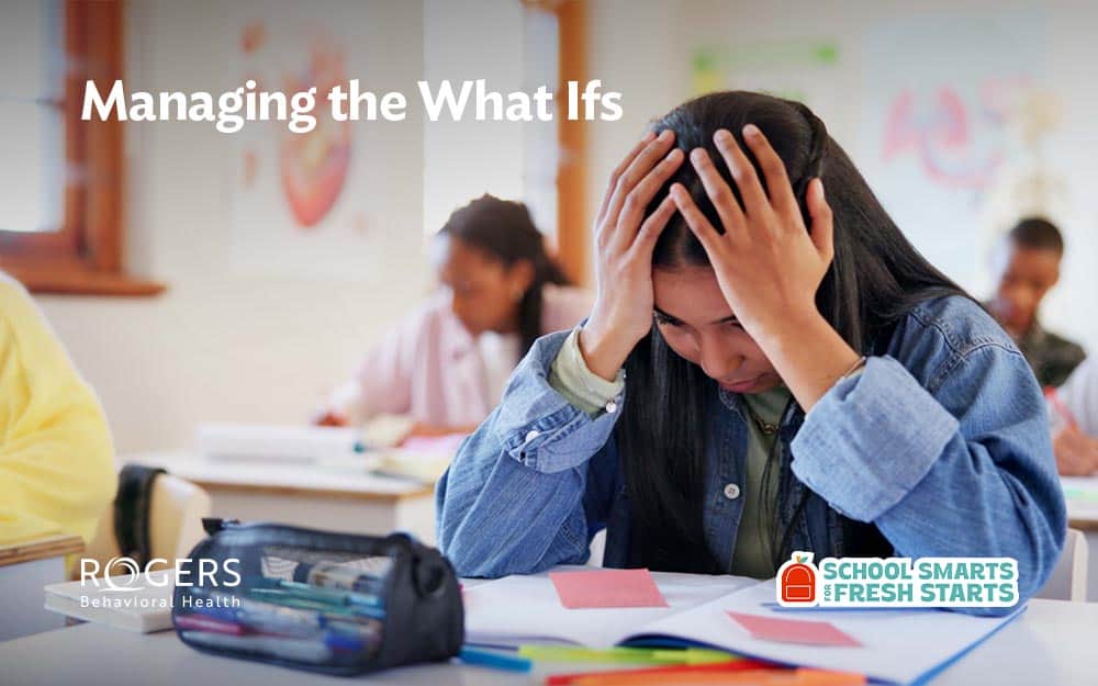 The image shows a classroom setting with several students seated at desks. The primary focus is on a student in the foreground with long dark hair, wearing a denim jacket. The student appears stressed, holding their head in their hands while looking down at a notebook. On the desk, a pencil case and some papers are visible. Other students are blurred in the background, engaged in their work. Transcribed Text: Managing the What Ifs
