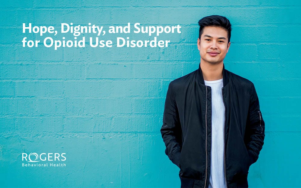 The image features a young adult standing against a vibrant turquoise brick wall. He is dressed in a black jacket over a white shirt, with his hands casually placed in his jacket pockets. The background is a consistent shade of turquoise, providing a clean and bold contrast to his dark attire. The left side of the image contains text in white, prominently displayed in the upper portion of the wall. Transcribed Text: Hope, Dignity, and Support for Opioid Use Disorder