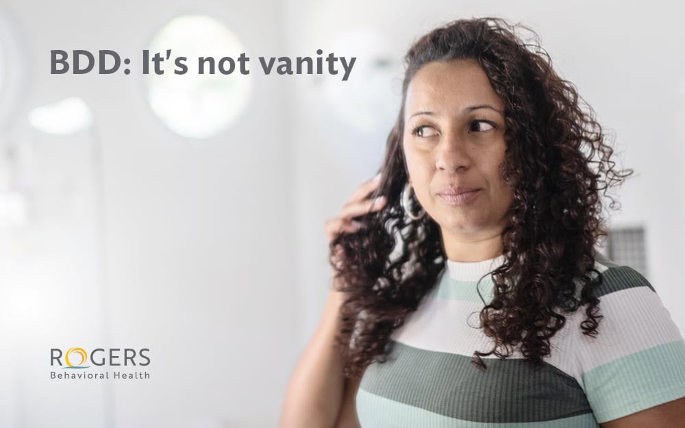 The image shows a woman with curly hair looking pensively to her left. She is wearing a striped shirt with shades of green and white. The background is softly blurred, with a white wall featuring a few circular light fixture