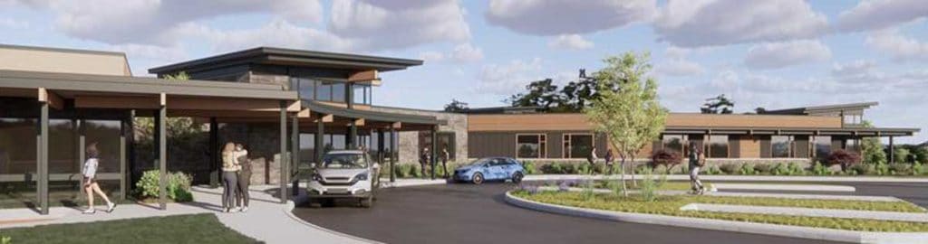 A modern building complex with large windows, stone, and wooden exterior. There are people walking, parked cars, and a roundabout with a small tree in front.